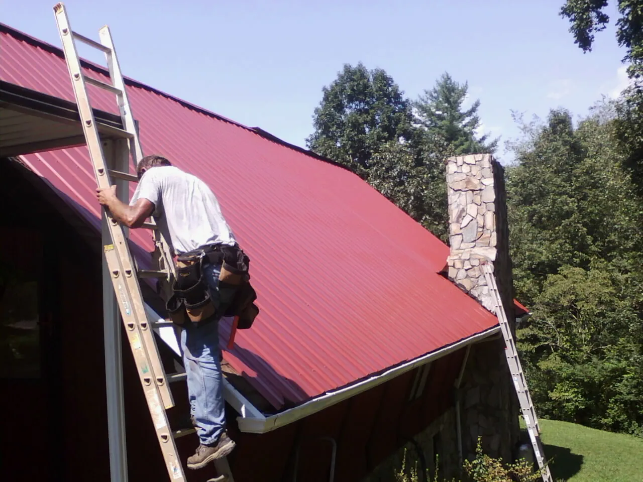 Roofer on a ladder beside a red metal roof and stone chimney — Soffit & Fascia Repair by Roofing Brothers in Matamoras